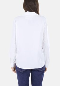 Camicia bianca a maniche lunghe con collo, tessuto liscio e polsini con bottoni, abbinata a jeans di denim blu. Vista posteriore mostra un orlo curvo.