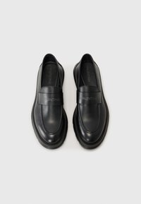 Black leather loafers feature a broad strap with "VALENTINO" embossed, rounded toe, and a subtle stitched design along the edges.