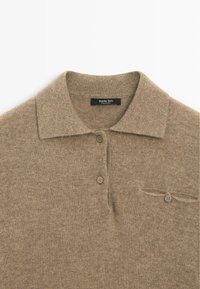 Polo beige lavorata a maglia con colletto, tre bottoni e una piccola tasca sul petto. Realizzata con tessuto morbido e strutturato. Etichetta del marchio visibile.