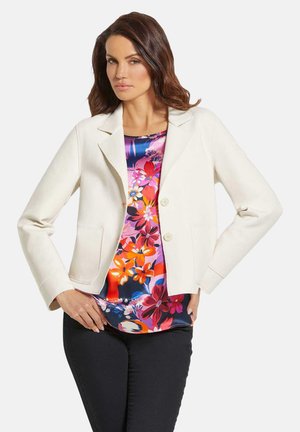 Weißer Blazer mit tailliertem Schnitt, ausgestattet mit zwei Fronttaschen und einem einzelnen Knopfverschluss, getragen über einem bunten, floral bedruckten Oberteil.