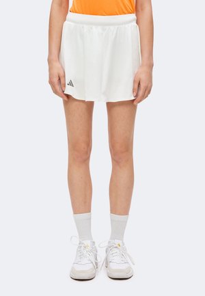 Personne portant une jupe de sport Adidas blanche, des chaussettes blanches et des baskets blanches, debout avec les mains détendues le long du corps.