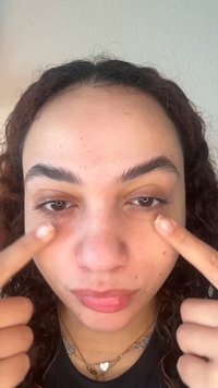 Person mit lockigem Haar zeigt auf dunkle Ringe unter beiden Augen, hebt Hautpflege- oder Make-up-Fokus hervor.