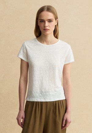 Giovane donna con capelli biondi lisci, indossa una semplice maglietta bianca a maniche corte e pantaloni marroni larghi su uno sfondo beige.