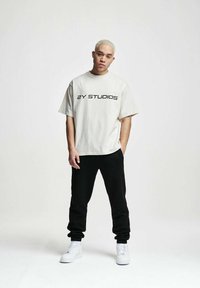 Valkoinen puuvillainen t-paita, jossa on teksti "2Y STUDIOS", yhdistettynä mustiin collegehousuihin. Mallilla on jalassaan valkoiset tennarit ja hän seisoo neutraalilla taustalla.