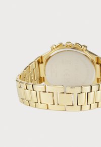 Montre en acier inoxydable doré avec un bracelet à maillons en bloc, un boîtier rond et une finition lisse et polie. La vue arrière montre les détails de la marque.
