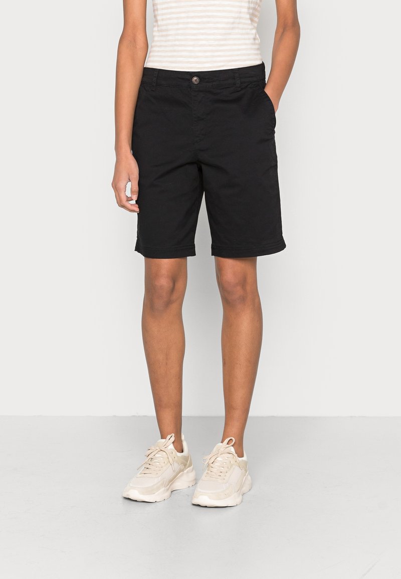 Osoba ve černých kraťasech ke kolenům, světle béžovém pruhovaném tílku bez rukávů a světle béžových teniskách stojící na jednobarevném pozadí.