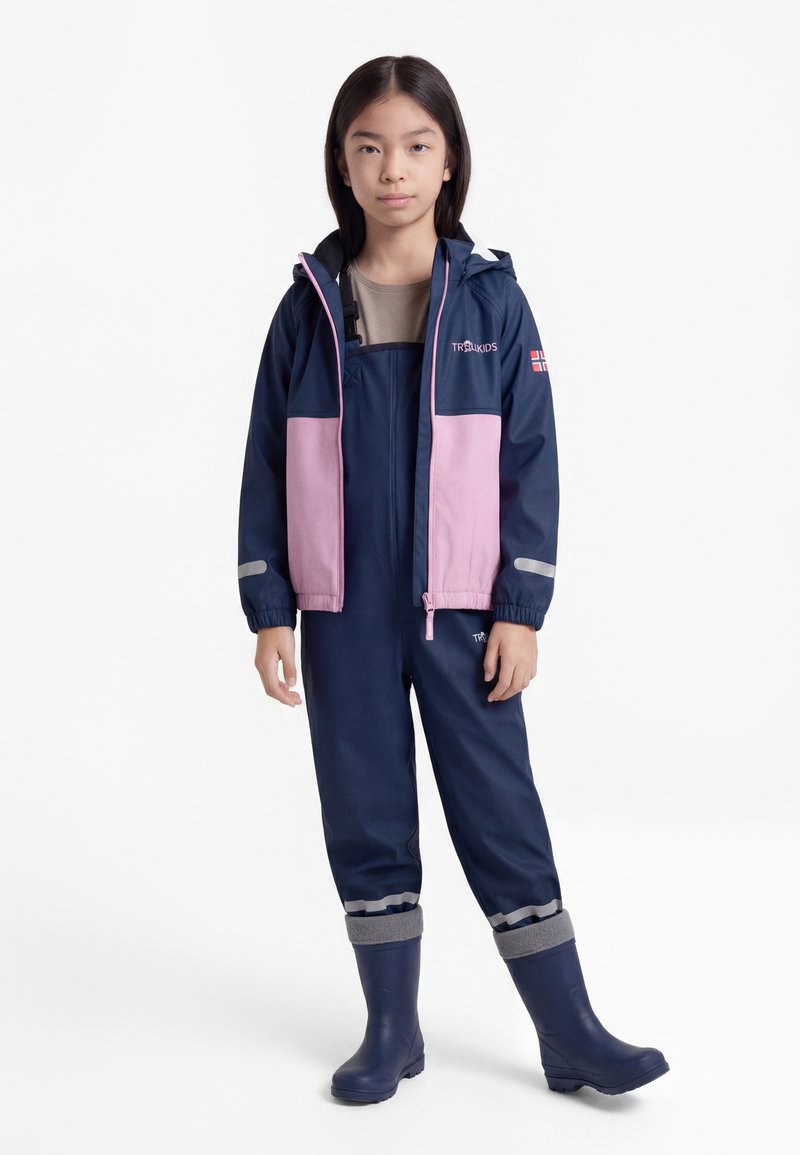 Παιδί που φοράει μπλε σκούρο και ροζ αδιάβροχο μπουφάν, μπλε σκούρα ολόσωμη φόρμα και λαστιχένιες μπότες, στέκεται σε λευκό φόντο.