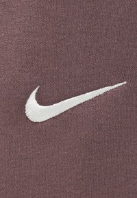 Baltas išsiuvinėtas Nike swoosh logotipas ant tekstūruoto violetinio audinio fono, rodančio detalius siuvinėjimo ir mezgimo medžiagos bruožus.