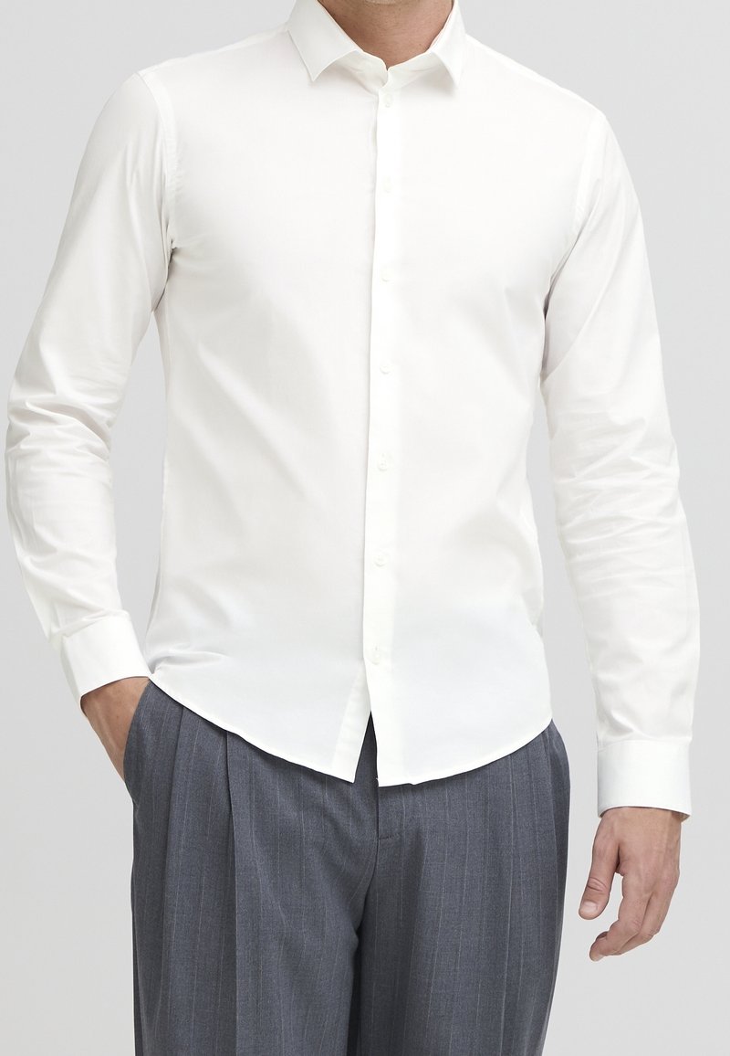 Homme portant une chemise blanche cintrée à manches longues rentrée dans un pantalon gris à fines rayures, une main dans la poche.