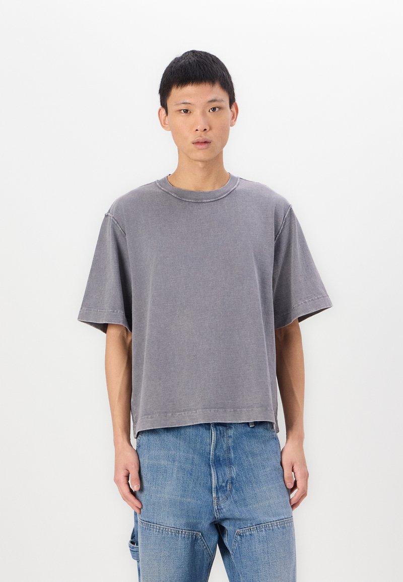 Tricou din bumbac gri, cu o croială lejeră, mâneci scurte, guler ribat și margine neterminată, asociat cu blugi din denim albastru deschis.