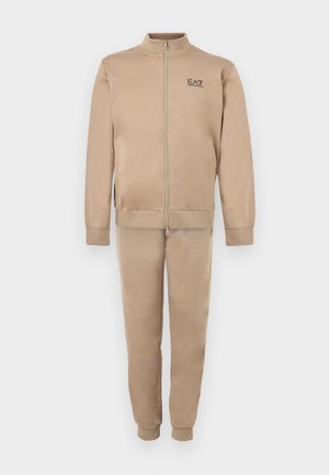 Trening bej din material fin, format dintr-o jachetă cu fermoar și guler înalt și pantaloni conici asortați, ambele cu branding discret.