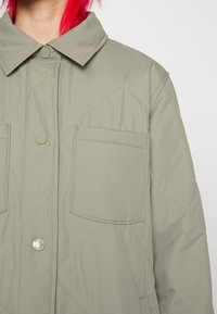 Kaki quiltad jacka med spetsig krage, två framfickor och tryckknappar. Tyget har en mjuk yta med matt finish.