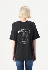 Juoda per didelė marškinėlių su baltu "COUTURE" grafiniais elementais, kuriuose pavaizduoti kaspinai ir tekstas "Belle Femme PARIS", dekoruotas studais, trumpomis rankovėmis.
