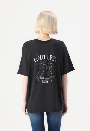 Zwarte oversized t-shirt met een witte "COUTURE" grafiek met strikken en de tekst "Belle Femme PARIS" versierd met studs, korte mouwen.
