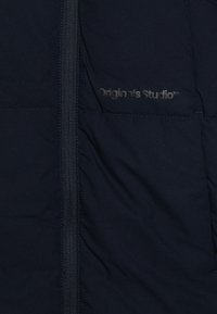 Mörkblå puffjacka med dragkedja, slät yta och "Originals Studio"-logotyp tryckt på framsidan. Lätt och isolerad design.