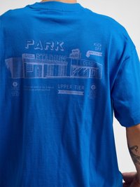 Tył niebieskiej koszulki z grafiką przedstawiającą fasadę stadionu, oznaczenia wejścia dla zawodników, salon, bramy A i B oraz tekst o sprzedaży biletów i dostępie.