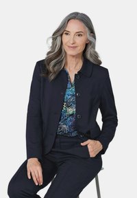 Navy blazer met ronde kraag, knoopsluiting en zakken, over een blouse met een veelkleurig bloempatroon. Model zit, met grijs haar.