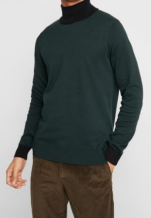 Homme portant un pull vert foncé avec un col roulé noir et des poignets, associé à un pantalon en velours côtelé marron, debout avec une main dans la poche.