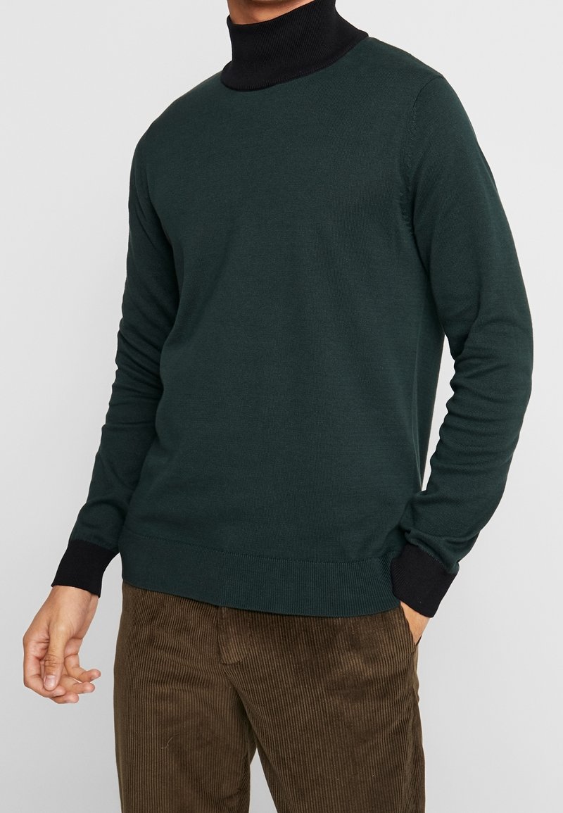 Homme portant un pull vert foncé avec un col roulé noir et des poignets, associé à un pantalon en velours côtelé marron, debout avec une main dans la poche.