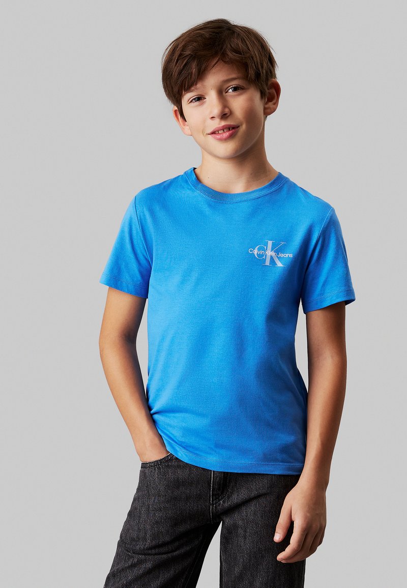 T-shirt di cotone blu brillante con maniche corte e scollo rotondo. Presenta un logo bianco sull'area del petto a sinistra. Indossato con jeans scuri.