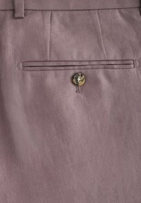 Gros plan de la poche arrière d'un pantalon en tissu mauve avec une fente horizontale et une fermeture par un bouton rond unique.
