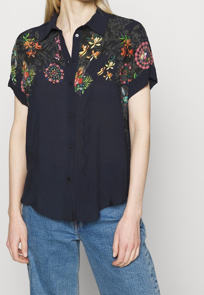 Chemise de couleur marine à manches courtes avec broderies florales en orange, rose et vert, dotée d'un devant boutonné et d'une coupe décontractée. Tissu texturé.