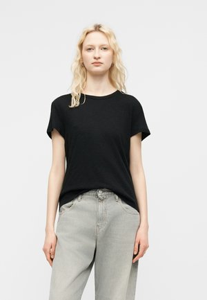 Femme blonde aux cheveux ondulés détachés, portant des boucles d'oreilles créoles en or, un t-shirt noir à manches courtes et un jean taille haute gris clair, debout devant un fond blanc.