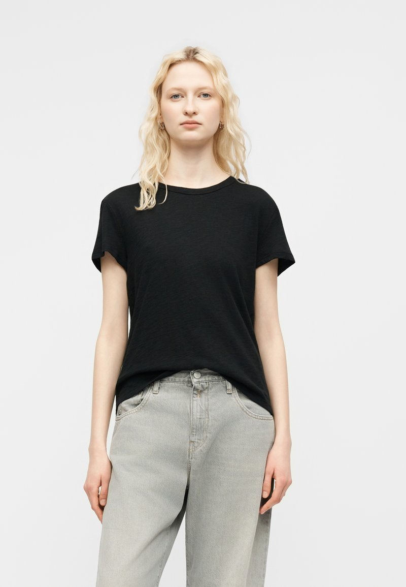 Femme blonde aux cheveux ondulés détachés, portant des boucles d'oreilles créoles en or, un t-shirt noir à manches courtes et un jean taille haute gris clair, debout devant un fond blanc.