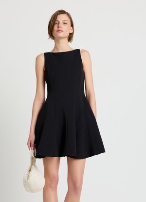Donna che indossa un vestito nero senza maniche aderente in vita e svasato, che tiene in mano una piccola borsa bianca testurizzata su sfondo neutro.