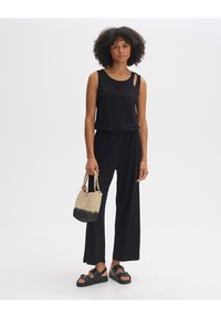 Schwarzer ärmelloser Jumpsuit mit Cut-Out-Detail, lockerer Passform und elastischem Bund. Das Model hält eine gewebte schwarze und beige Tasche und trägt schwarze Sandalen.