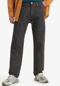 Man draagt zwarte spijkerbroek, witte sneakers, een blauw shirt en een mosterdgele blouse met één hand in de zak.