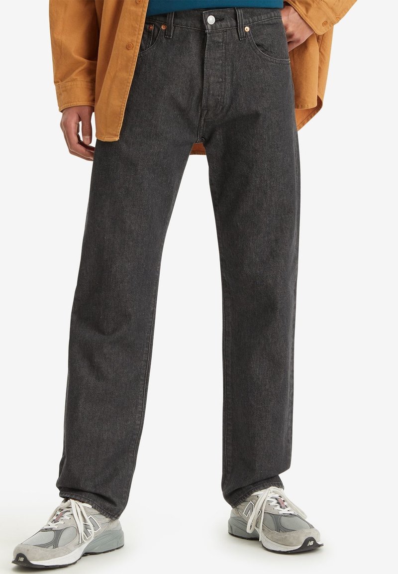 Man draagt zwarte spijkerbroek, witte sneakers, een blauw shirt en een mosterdgele blouse met één hand in de zak.