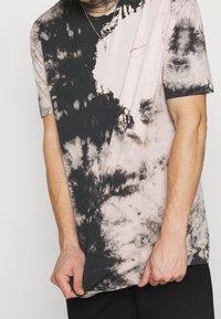 Camiseta de manga corta con teñido anudado en beige y negro con patrones en espiral. Cuello redondo, tejido suave y un detalle de logo mínimo en el pecho.