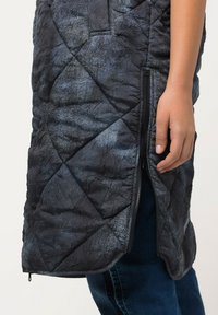 Genähtes Weste in Schwarz mit strukturierter Oberfläche, ausgestattet mit einem seitlichen Reißverschluss. Das Design zeigt ein geometrisches Muster und glatte Kanten.