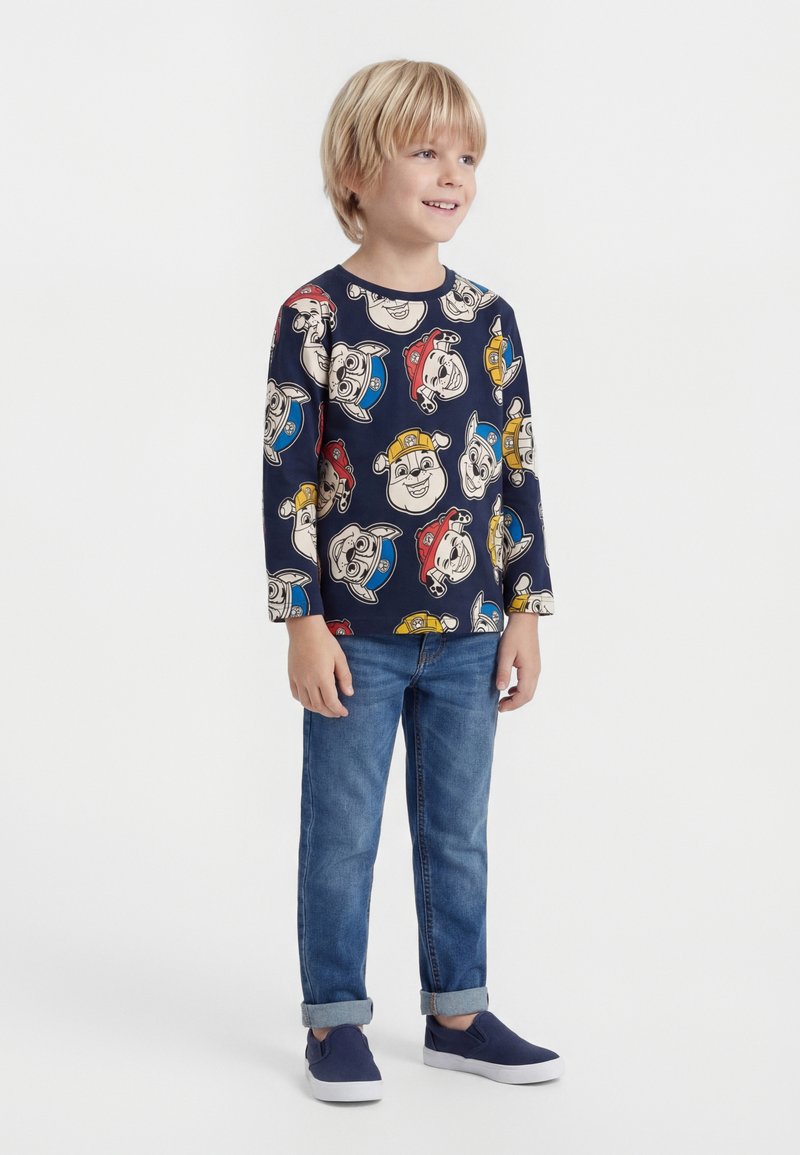 Svetlovlas fant nasmejan, oblečen v dolg rokav mornarsko modre barve z barvitimi risankarskimi obrazki psov, modre zvite kavbojke in mornarsko modre čevlje za natikanje.