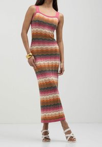 Robe maxi en tricot avec un motif en zigzag dans des tons de rose, orange, blanc et vert. Dotée de larges bretelles roses et d'une silhouette ajustée. Le modèle porte des sandales blanches.