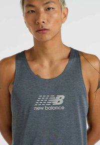 Grijze sportieve tanktop met een gestructureerd patroon. Heeft een wit "New Balance" logo op de voorkant. Mouwloos ontwerp, ontspannen pasvorm.