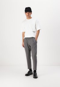 White short-sleeve t-shirt with a pocket and logo, paired with gray tapered trousers and black shoes. Black cap with logo detail.