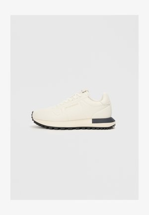 Ljust beige sneaker med en slät syntetisk överdel, texturerade detaljer, snörning och en svart gummisula för bra grepp.