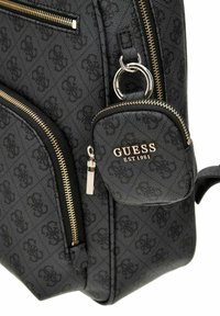Sac texturé noir avec des fermetures éclair dorées et une petite pochette détachable. Motif monogrammé embossé sur la surface. Logo de la marque visible.