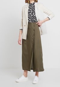 Olivgrüner, fließender, knöchellanger Rock mit Knöpfen vorne; kombiniert mit einem floralen schwarzen Oberteil und einem hellen Blazer; weiße Turnschuhe.