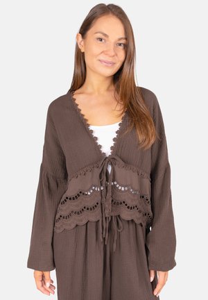 Jeune femme portant un haut marron à manches longues avec un nœud à l'avant et des détails en dentelle festonnée, sur un sous-vêtement blanc, posant devant un fond uni.