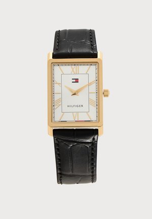 Orologio rettangolare in oro con quadrante bianco, numeri romani, lancette dorate, cinturino in pelle nera e logo e nome Tommy Hilfiger sul quadrante.