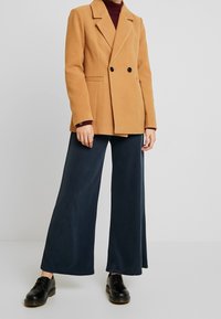 Brun dubbelknäppt blazer med slag, i kombination med vida marinblå byxor och svarta snörskor. Slät textur, skräddarsydd passform.