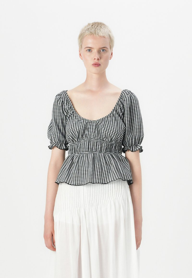 Top peplum a quadretti bianchi e neri con maniche a palloncino corte e scollatura profonda, abbinato a una gonna bianca con pieghe. Tessuto crisp e strutturato.