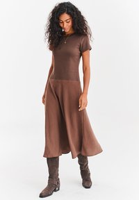 Brązowa sukienka z krótkimi rękawami, dopasowanym gorsetem i rozkloszowanym dołem. Zestawiona z brązowymi zamszowymi kozakami za kolano. Prosty wzór i strukturalna tkanina.