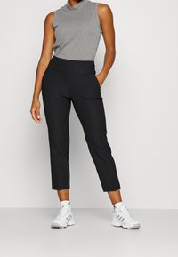 Černé zkrácené kalhoty s hladkým povrchem, obsahující boční kapsy a vysoký pas, v kombinaci se šedým bezrukávovým topem a bíl ými teniskami.