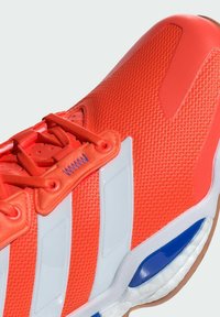 Červená sportovní bota s texturovaným svrškem, bílými pruhy a oranžovými tkaničkami. Má modro-bílou podrážku navrženou pro odpružení a podporu.