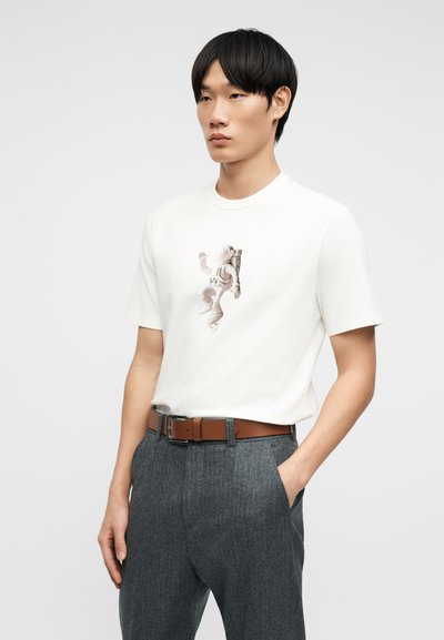 Νέος άνδρας που φοράει λευκό μπλουζάκι με γραφικό αστροναύτη, γκρι παντελόνια και καφέ ζώνη, στέκεται με το ένα χέρι στην τσέπη.