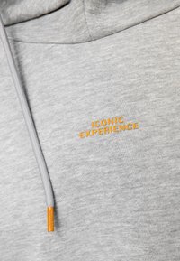 Tissu de sweat à capuche gris clair avec gros plan sur le texte orange "EXPÉRIENCE ICONIQUE" et embout de cordon assorti orange.